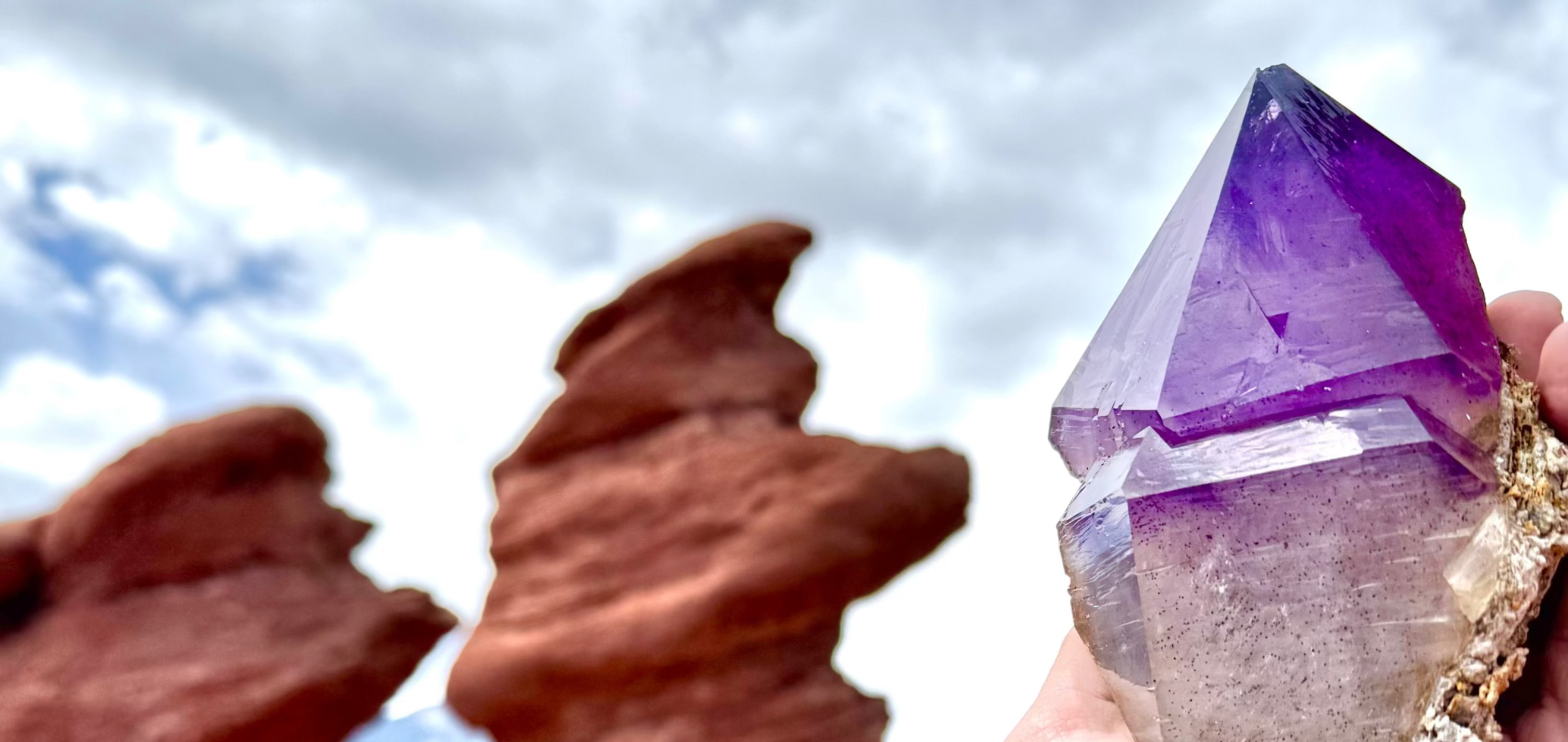 Amethyst crystal held against a natural background with red rock formations.